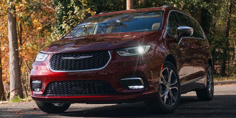 Red minivan parked on a paved road in a wooded area, shown from a front three-quarter angle with headlights illuminated