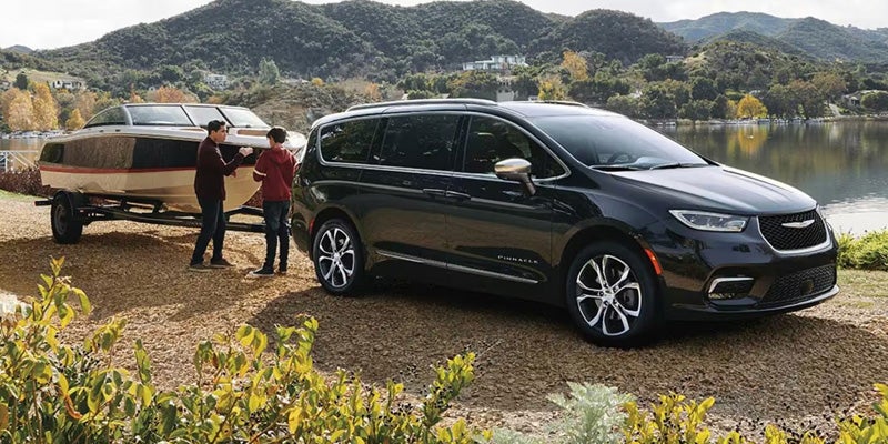 Black minivan parked beside a lake with a boat trailer attached, set against a backdrop of rolling hills and trees