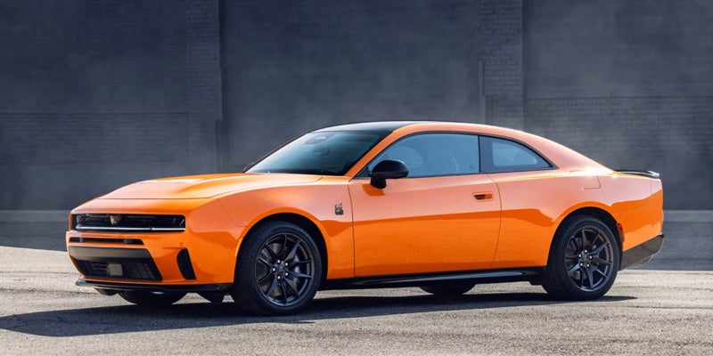 Orange two-door muscle car shown in side profile, parked on a paved surface in front of a dark gray wall during daylight