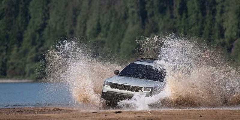 Silver SUV driving through shallow water near a lakeshore, creating large splashes on both sides of the vehicle