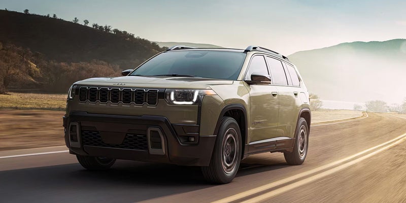 Olive-green SUV driving on a paved road through a scenic landscape with hills and morning haze in the background