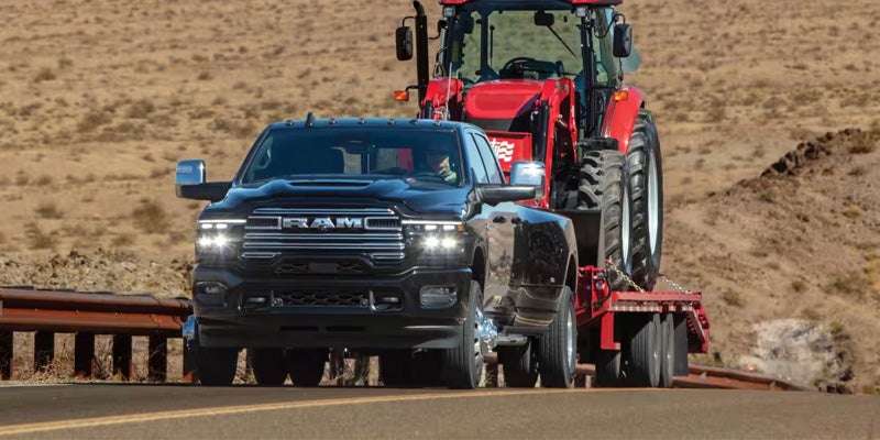 Black heavy-duty pickup truck towing a red tractor on a trailer along a paved road through a desert landscape
